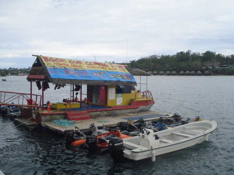 A floating pontoon and three boats belonging to U Power Adventures on the Port Vila waterfront. There is a big banner covering the roof and advertising the business. Lifejackets are hanging up. The sky is overcast