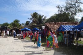 Stalls along a beach in Vanuatu. The stalls are mostly palm thatch and are selling colourful clothing. There are palm trees behind