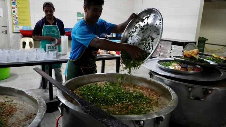 A worker tips spring onions and fried shallots into a giant vat of rice porridge. The surface of the porridge is covered in green.