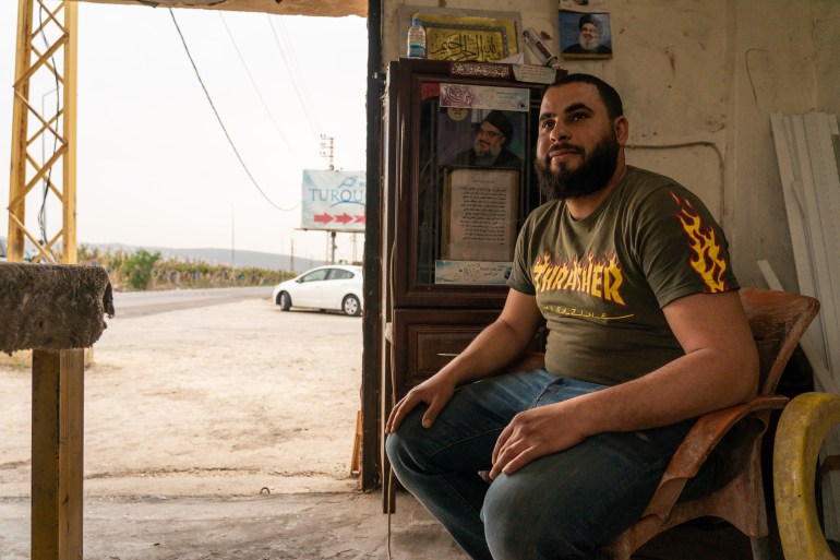 Man sits in a room next to an open door with a small picture of Hezbollah leader Hassan Nasrallah stuck on a wall in the background