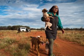 Gloria Morales with some of the dogs she cares for in Australia's Northern Territory aboriginal community of Yuendumu. She is walking on a dirt road with a puppy in her arms while other dogs follow along.
