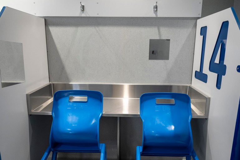 The cubicle has a steel table and two blue chairs. There is a large number 14 on the right-hand side of the partition wall.