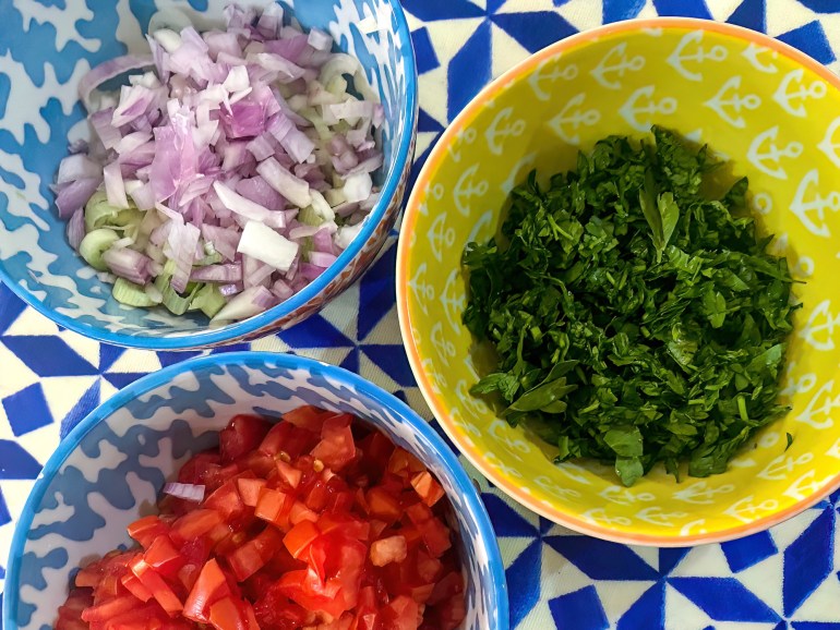 a bowl of chopped tomato, another of chopped onion, and a third of chopped parslet on a blue and white surface