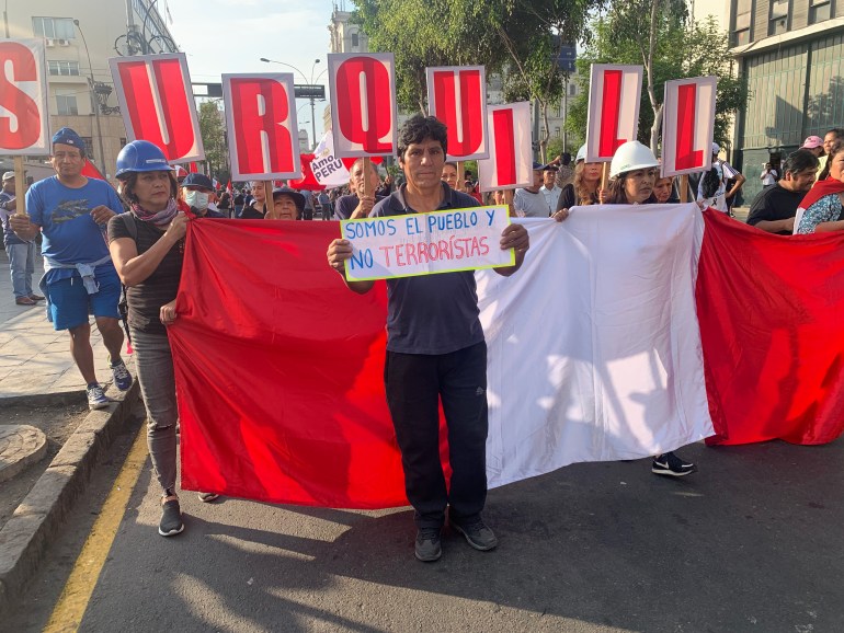 A man holds a small handwritten sign in front of a line of protesters carrying a banner with the colours of the Peruvian flag