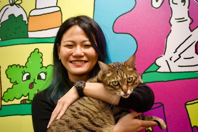 Zoe is smiling, wearing a black, long-sleeve top and black watch and holding a cat. She is standing against a colourful mural.
