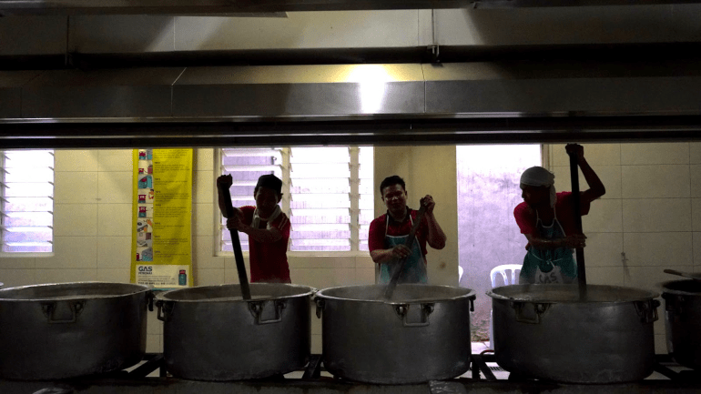 Three men stirring giant pots of rice porridge in the Kampung Baru mosque's kitchen. They are using giant paddles to stir the mixtire and are silhouetted against the windows 