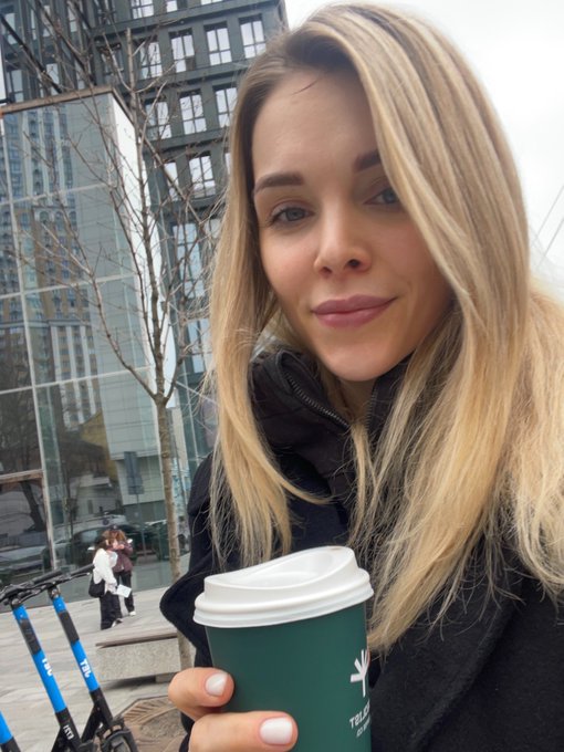 A close-up of Victoria, with straight blonde hair, wearing a black coat and holding a green takeaway coffee.