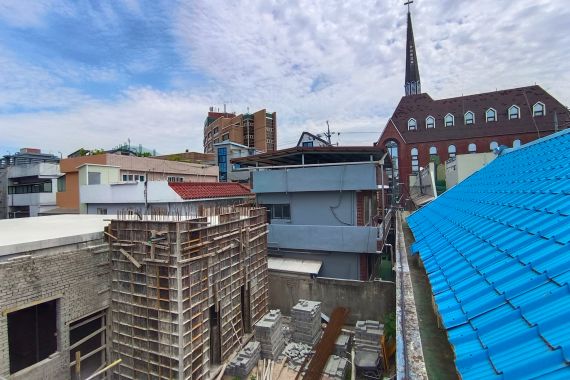 View of roof tops from the temporary prayer room at the mosque showing the construction site. There is a church with slender spire and a cross on top 30 metres behind, and lots of tightly packed low-rise buildings.