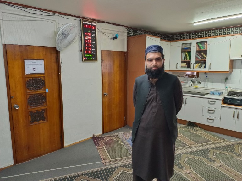 Muaz Razaq inside the temporary prayer hall. There are kitchen cabinets behind him and a cooker, a fan and an LED clock on the wall. He is wearing traditional dress in black and brown., and has a bushy black beard.