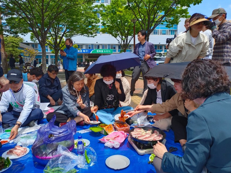 Anti-mosque protesters sit on a blue tarpaulin as they barbecue pork after a protest outside the Buk district office. Some of the women are holding umbrellas and wearing face masks. One is helping herself to kimchi.