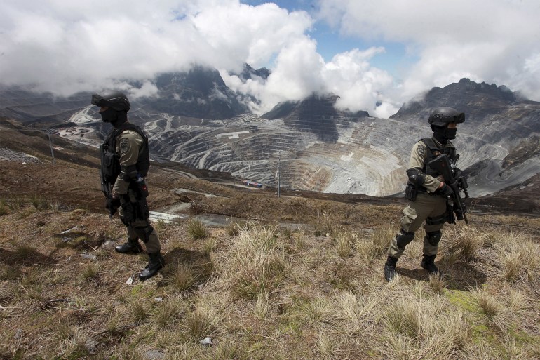 Two Indonesian police officers stand guard at the Grasberg mine. They are armed and in military-style outfits. The open-pit mine, a vast hole carved out of the mountains, is behind them.