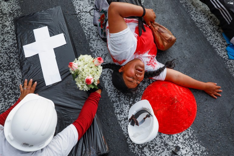 Protesters lie on a road splattered in blood-like paint. A black coffin sits next to them, and another person leans over to deliver flowers.