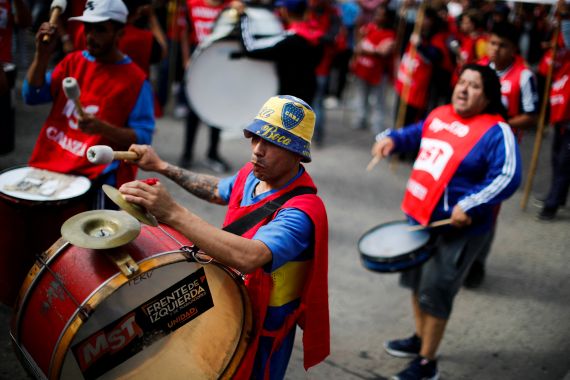 A man in a yellow and blue bucket hat plays a drum strapped to his chest. Other protesters can be seen around him, also playing drums.