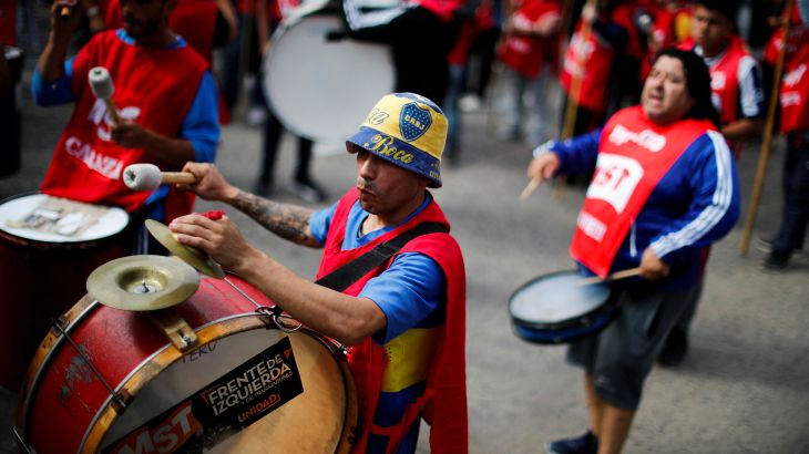 A man in a yellow and blue bucket hat plays a drum strapped to his chest. Other protesters can be seen around him, also playing drums.