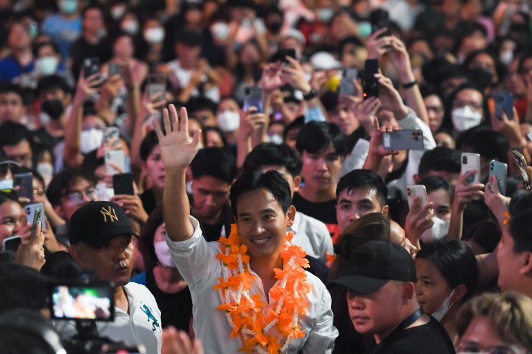 Pita Limjaroenrat, Move Forward Party's leader, at a party campaign event. He has an orange garland around his neck and is waving and smiling, There is a crowd behind him.