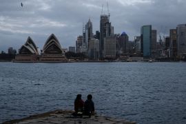 A nightfall skyline view of Sydney Harbour with the Sydney Opera House and the central business district om the background. Two people are silhouetted sitting on an outcrop in the foreground.