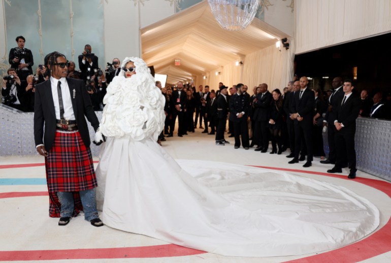 Rihanna and her partner ASAP Rocky pose for photographers at the Met Gala. She is wearing a white dress with a long train. He is in a tartan kilt with white shirt and black jacket.