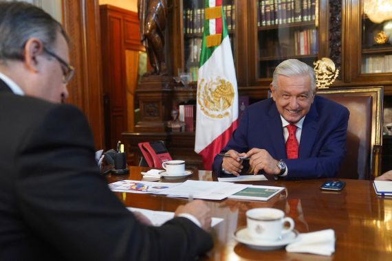 Mexico's President Andres Manuel Lopez Obrador and Mexico's Foreign Secretary Marcelo Ebrard hold a call at the National Palace in Mexico City. Lopez Obrador is seen on the far side of the wooden table, in front of a Mexican flag and a glass-shielded book case.