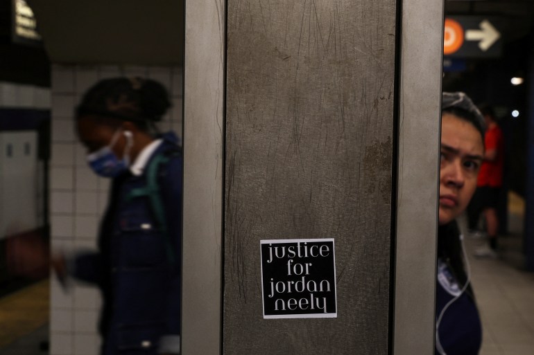 A wall in a New York City subway station pole has a sticker on it that reads: "Justice for Jordan Neely". A woman looks at the camera from halfway behind the pole and on the other side is a blurry image of another woman with a mask on.