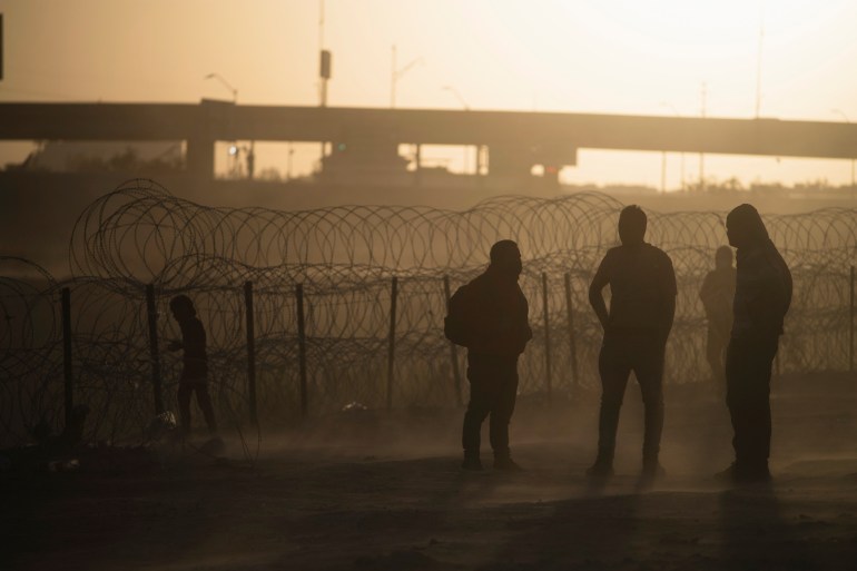 Asylum seekers stand on a dusty field near barbed wire at the US-Mexico border