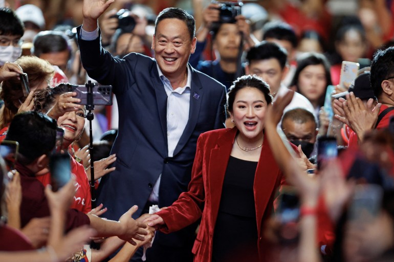 Paetongtarn Shinawatra, daughter of former Prime Minister Thaksin Shinawatra, is wearing a velvet crimson jacket, a gold necklace and a black top. She is walking through crowds whose hands are outstretched towards her, some holding up their cameras. She is smiling and one of her hands is loosely holding a hand in the crowd that she looks about to drop before the next one.