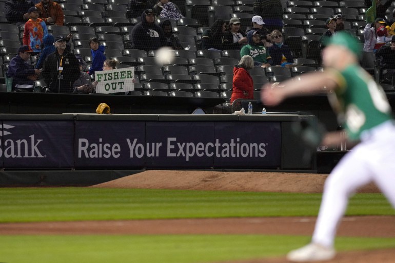 A Oakland Athletics fan holds up a sign directed at owner John Fisher as relief pitcher Sam Moll (foreground) warms up