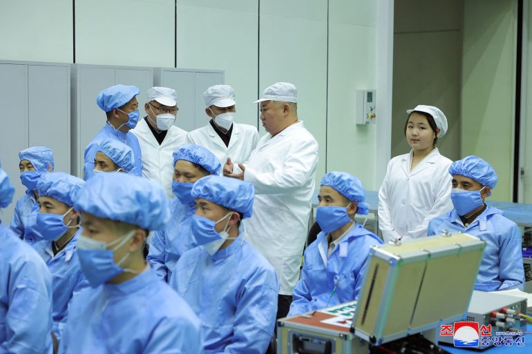Kim Jong Un in a white lab coat and hat talking to officials from the satellite development committee. Some are in white lab coats and others in blue. Kim's daughter Kim Ju Ae is behind him and slightly to one side. Other workers are sitting in the front. Everyone is wearing a face mask except the Kims.