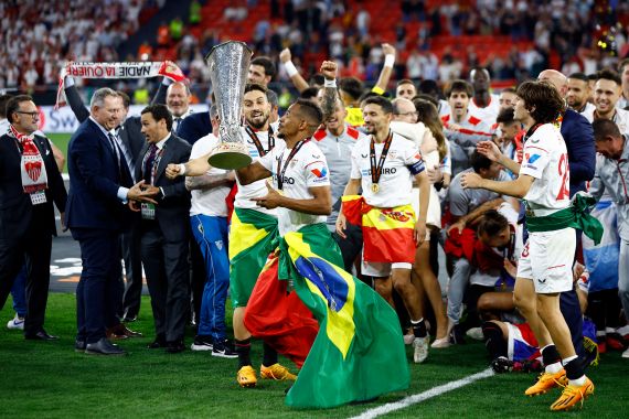 A football player, dressed in a white kit and waving a Brazilian flag, lifts aloft the Europa league trophy, while his teammates celebrate.