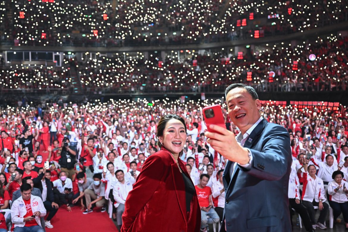 Paetongtarn Shinawatra (L) and Srettha Thavisin (R) take a selfie photo on stage at the party's final campaign rally. There is a sea of supporters in red. They have tuned on the lights on their smartphones.