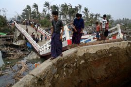 People stand on a broken bridge following Cyclone Mocha. The bridge is broken in two. There are people standing on the other bank and boats in the water