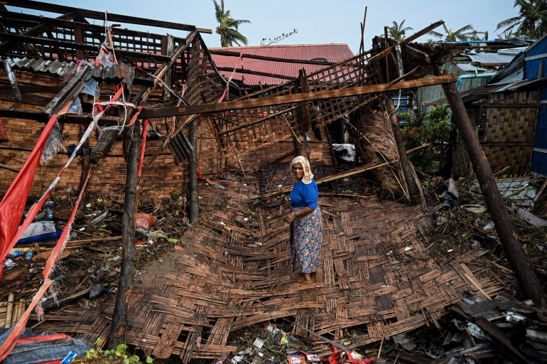 A Rohingya woman standing on what remains of her house near Sittwe after Cyclone Mocha. She is standing on the bamboo screens that were once the walls of the house. The wooden structure is twisted and broken.