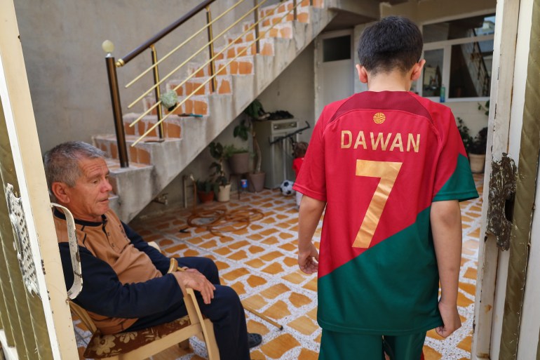 A photo of Dawan walking into the family home in Erbil, wearing a red and green football shirt as his father sitting next to him looks on.