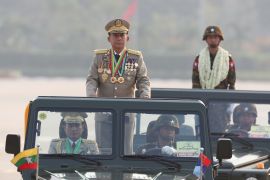 Senior General Min Aung Hlaing inspecting troops from an open top jeep. He is wearing lots of medals and standing to attention.