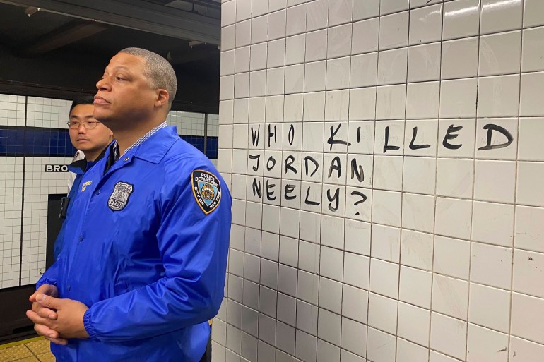 A police officer in a blue shirt stands in front of a white tiled wall, inscribed with the message: Who killed Jordan Neely?