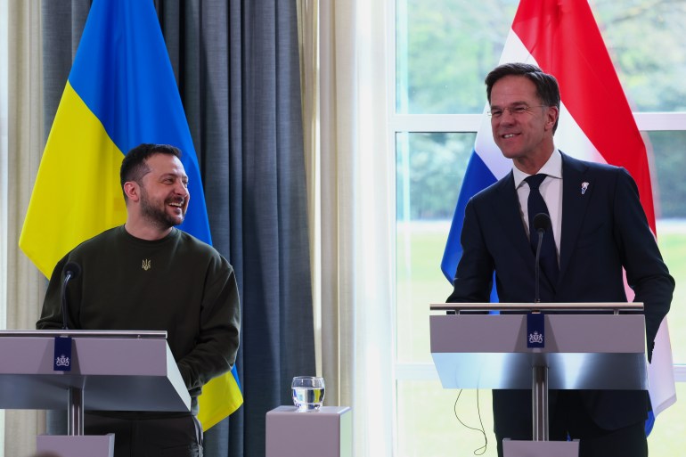 Zelenskyy behind a podium is looking to his left at Mark Rutte and laughing. He is wearing his customary military jersey and the Ukrainian flag stands behind him. Rutte is also smiling but looking forward from behind his podium, with the Dutch flag behind him.
