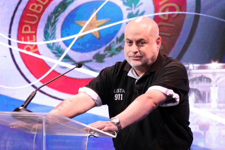 A bald man in a black shirt stands at a glass podium, as a graphic of the Paraguay flag is seen behind him