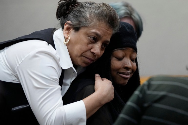 A woman leans over to hug a second woman from behind as she cries in the courtroom