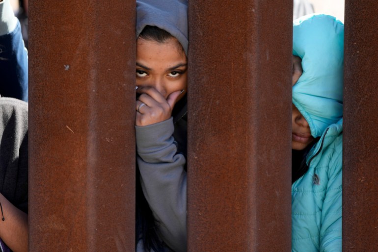 Migrants waiting to apply for asylum between two border walls look through the wall