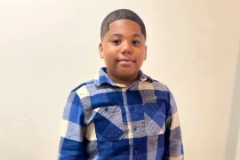 A pre-teen boy stands against a beige background in a blue plaid shirt.