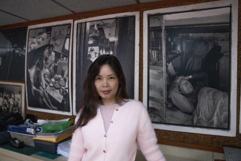 Sze Lai-shan standing in front of large printouts of black and white photographs showing dire living conditions. She is looking confidently at the camera.