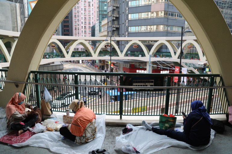 Domestic workers relaxing on an elevated walkway in Hong Kong on a day off. They are sitting on mats, looking at their phones and chatting to each other.