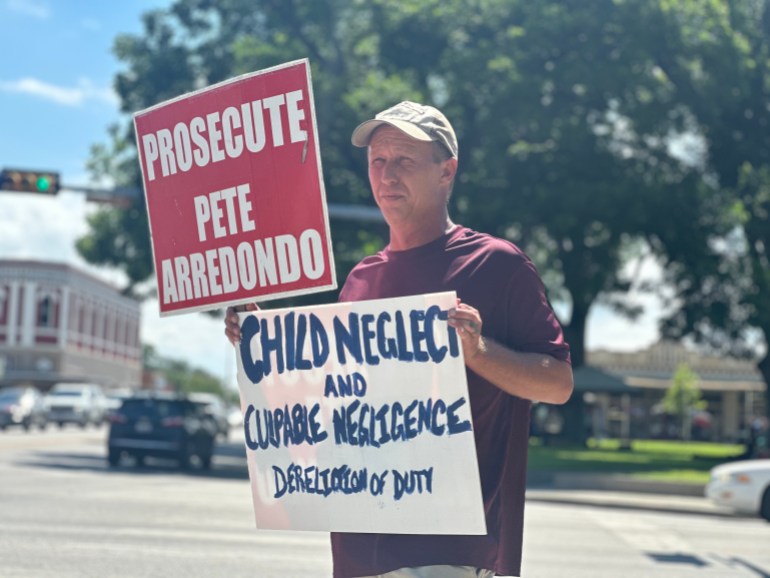 Uvalde resident Mike Brown holding two placards. One says Prosecute Pete Addedondo. The other Child Neglect and Culpable Negligence, Dereliction of Duty