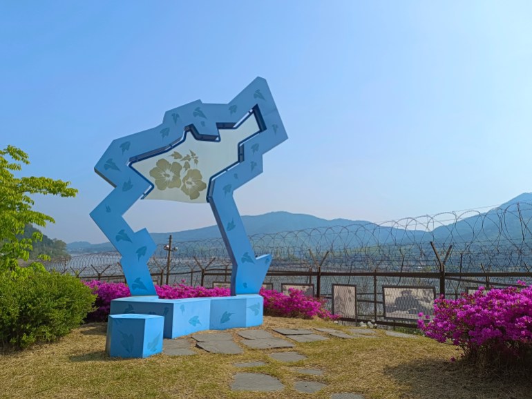 A blue sculpture to veterans of the Korean war in the DMZ. There is pink bougainvillea and mountains behind.