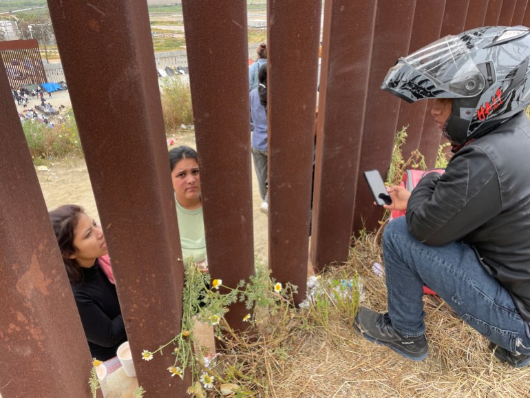 A man in a motorcycle helmet leans down with a phone to take a food order from women on the other side of a border wall