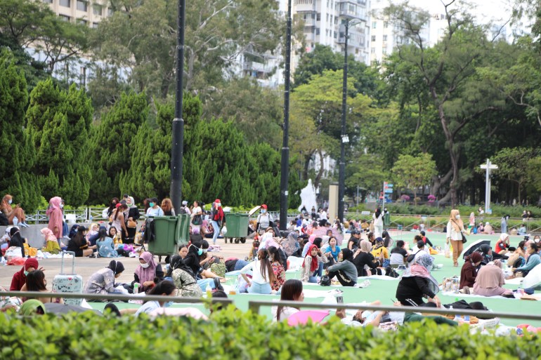 Indonesian domestic workers hang out in Victoria Park on their day off. They are sitting on ht ground. There are trees and skyscrapers behind them.