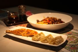 A rectangular plate carries mantu dumplings and half of a circular gosh naan pastry. Behind, a bowl of laghman noodles sits.