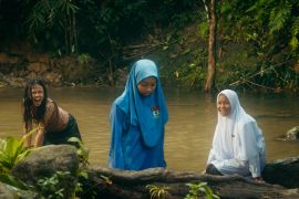 A still from Tiger Stripes showing three girls standing in a river in Malaysia. Two are in school uniform and wearing head scarves. The third is behind them crouching down. She's wearing a black t-shirt and her hair is loose and messy. The river water is brown ad there are trees behind.
