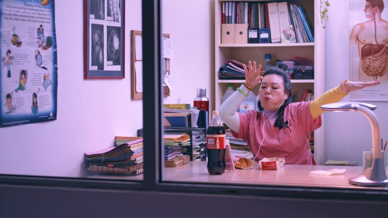 A still from Vinegar Baths showing a woman sitting at an office desk in what looks like a clinic. There are diagrams of anatomy on the wall and a book case behind her with files. She has a bottle of Coke and some french fries on the desk. She has her earphones on and is moving her arms to what is playing on her phone.