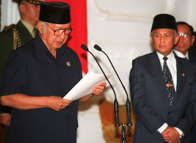 Suharto announces his resignation. His vice president BJ Habibie is standing nearby, and there is an Indonesian flag behind. Suharto is reading from a sheet of paper