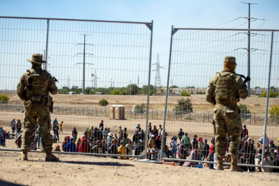 Armed soldiers watch migrants through a fence